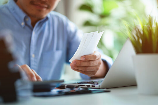 A man is sitting at a desk with a laptop and a stack of receipts. He is holding a receipt in his hand and he is organizing his finances