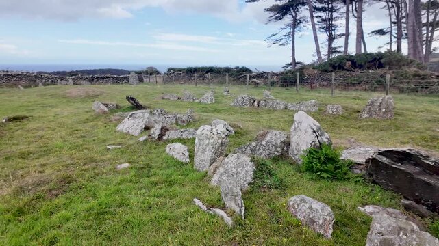 Ancient stone tomb remains scattered across a grassy field with coastal views and trees in the Isle of Man.. Pan Left