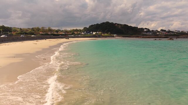 Low drone footage of shoreline at the stunning Cobo Bay Guernsey Channel Islands with golden sand clear turquoise water and lapping waves in the sunshine with dramatic sky