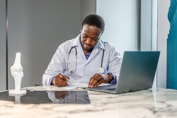 Young doctor man working at office workplace, reading medical record, paper document at laptop, reviewing patient history, sitting at table with laptop computer, working in clinic office