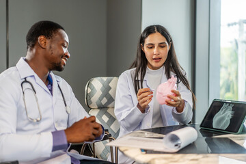 Two cardiologists engage in a detailed discussion on heart disease treatment, using an anatomical model to illustrate their points while consulting with a medical intern in a clinic, share knowledge