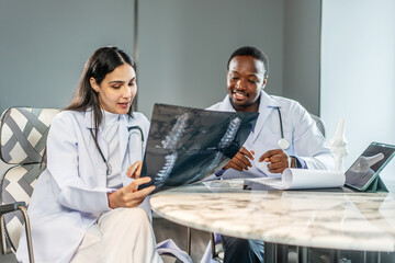 Two diverse doctors collaborate in a medical hospital, examining test results and discussing X-ray scans and use tablet computer. They focus on patient health and treatment strategies.