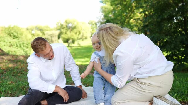 Joyful family moment in park: baby takes first tentative steps with parents' support, and happy mother kisses sweet baby. Family enjoys time outdoors