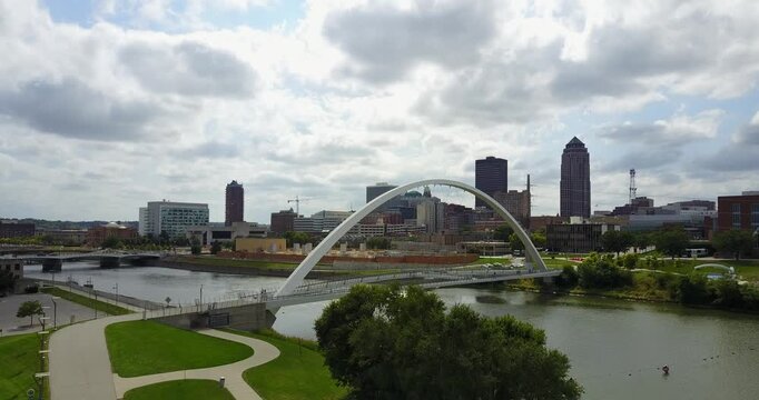 Aerial drone video of the river water going under a white arch bridge as the camera moves up and right to reveal the city skyline buildings of Des Moines, Iowa, USA.