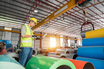 An Industrial engineer in safety gear uses a laptop to check inventory data among stacks of colorful metal coil rolls in a large factory warehouse.