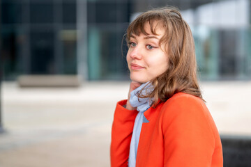Obraz premium Young woman in orange coat with blue scarf gazes thoughtfully in an urban setting on a sunny day