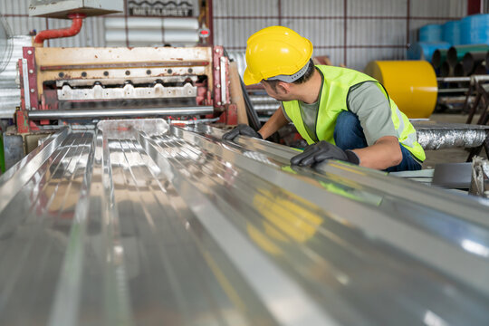 A Worker in safety gear inspecting the quality of a corrugated metal sheet (roofing panel) as it is produced by a roll forming machine in a factory.
