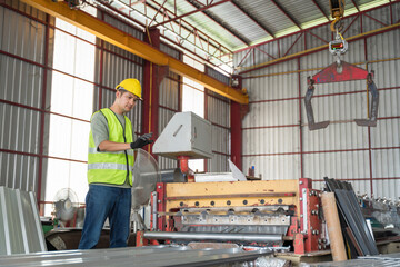 A factory worker in safety gear checking the control box of a roll forming machine used to produce metal sheets in a bright industrial warehouse.