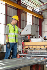 Asian technician in safety vest and hard hat adjusting the control panel of a metal sheet roll forming machine in a large factory workshop.