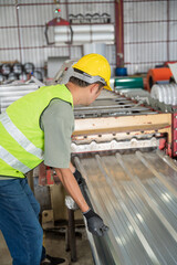 Asian Worker in safety gear inspecting and guiding a freshly cut corrugated metal sheet as it moves along the roll forming machine in a factory.