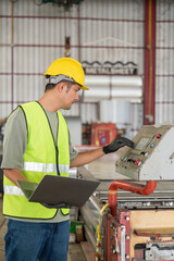 Asian engineer using a laptop and operating the control panel of a heavy roll forming machine in a metal sheet manufacturing factory.