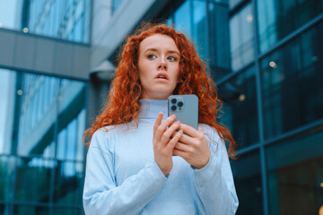Young woman with red curly hair looks surprised while using her smartphone outside modern building