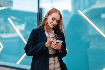Young woman using smartphone while standing near modern glass building in urban setting during daytime