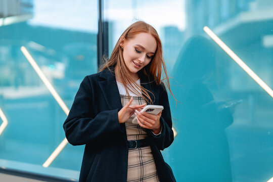 Young woman using smartphone while standing near modern glass building in urban setting during daytime
