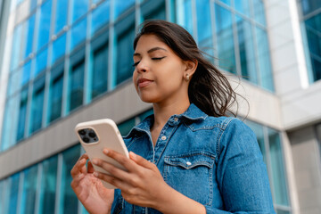 Young woman interacting with smartphone in front of modern glass building during sunny day