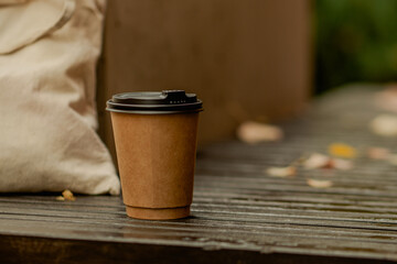 Paper coffee cup and eco bag on a bench in autumn with orange fall leaves in the background.