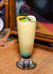 A vibrant cocktail in a tall glass, garnished with fresh fruit, sits on a wooden table, beckoning the viewer with its refreshing appearance.