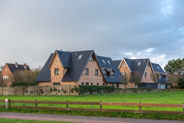 Fototapeta premium Residential brick houses with dark roofs. Modern brick houses with steep dark roofs in a quiet suburban neighborhood. The view shows fenced gardens and green lawns under a cloudy sky.