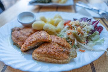 Plate of fried fish fillets served with boiled potatoes and fresh salad. A small bowl of sauce accompanies the hearty meal on a wooden table.