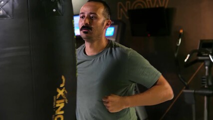 Amateur man with mustache awkwardly hitting a punching bag. Man in his 30s with no experience trying to box and kick a heavy bag in a gym. 4K.
