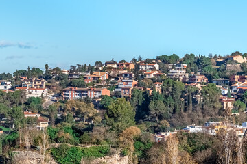 Fototapeta premium Mediterranean hillside community in Girona with modern homes among pine trees, terracotta roofs, and lush greenery under clear blue skies, showcasing elegant suburban living. Girona, Spain