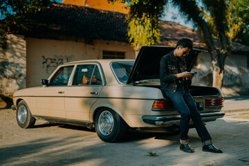 Stylish Asian man in black denim jacket leaning on vintage car with open trunk while using smartphone, cinematic retro lifestyle scene under warm afternoon light. © Vlafy Studio