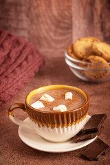 Cozy hot chocolate drink or homemade sweet cocoa garnished with marshmallows served in ceramic cup on plate on dark brown table with cookies and warm knitted sweater or blanket as breakfast dessert
