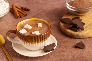 Sweet hot chocolate drink or homemade cocoa served in cup garnished with marshmallows served on dark brown rustic table with cinnamon spice, ingredients and spoon as dessert for cozy winter holidays