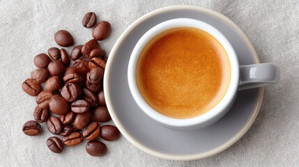 Fototapeta premium Overhead View Of A Ceramic Cup Filled With Rich Espresso Coffee With A Frothy Crema Next To A Pile Of Roasted Coffee Beans On A Textured Neutral Fabric Background In Soft Natural Light