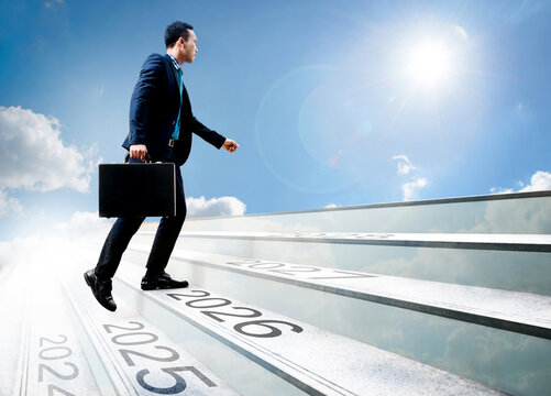 Happy new year 2026. Young Asian businessman holding black briefcase walking up the stair with sunlight sky background. 2025 2026 2027 on the upward staircase, concept of starting in the new year.