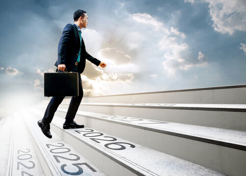 Happy new year 2026. Young Asian businessman holding black briefcase walking up the stair with sunlight sky background. 2025 2026 2027 on the upward staircase, concept of starting in the new year.