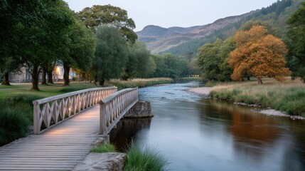 Fototapeta premium Wooden Footbridge Illuminated by Warm Lights Over a Shallow River in a Scenic Landscape With Mountains and Lush Green Trees During Twilight