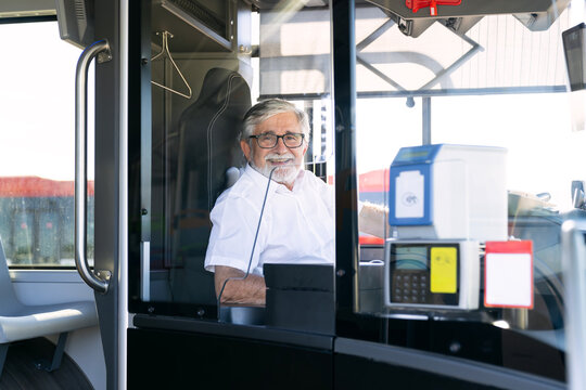 Mature man driving a city bus, happily looking at the camera from his driver's cabin, representing public transportation