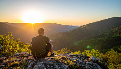 Fototapeta premium A young man sitting on top cliff in spring mountains at sunrise, back view, beauty of nature