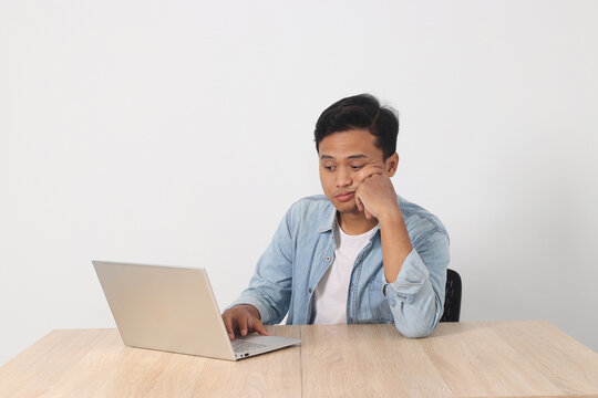 Young Asian man feeling bored and tired while working on his laptop with a frustrated expression, resting his head on one hand. Concept of work fatigue, burnout, or lack of motivation