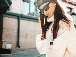Portrait of beautiful smiling model. Woman dressed in warm hipster white sweater and beanie. She posing in the street at sunset in sunglasses