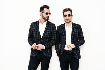 Portrait of two handsome confident stylish hipster lambersexual models. Sexy modern men dressed in black elegant suit. Fashion male posing in studio near white wall. In sunglasses