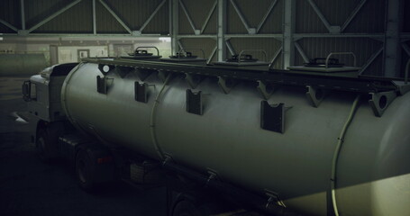 A large tanker truck is parked inside a spacious warehouse. The setting is dimly lit, emphasizing the tankers robust design while shadows play on the walls.