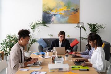 Four diverse women are working diligently on their laptops at a modern office table. They are focused on their tasks in a collaborative workspace, surrounded by plants and art.