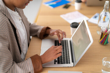 A person with dark skin types on a laptop at a wooden table. They are working in a bright office setting, surrounded by various supplies like pens and beverages.