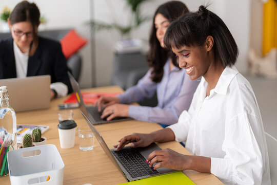 Three diverse women collaborate in a modern office, each focused on their laptops. The smiling Black woman in the foreground actively types, alongside her concentrating colleagues.