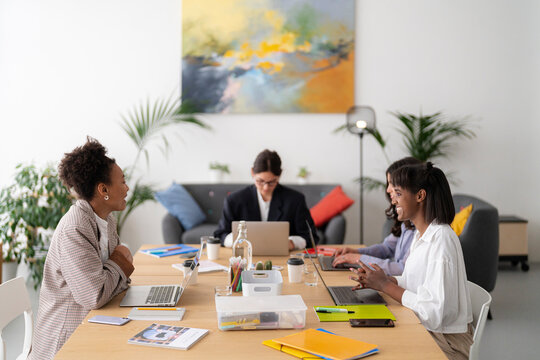 Diverse professional women collaborate in a bright office. They engage in conversation and work on laptops at a shared table, promoting teamwork and innovation.