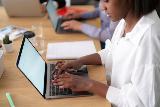 A young Black woman types on a laptop at a wooden desk, holding a pen. She is focused on her work in a modern office environment. Another person works in the background.