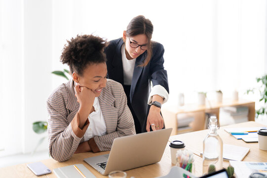 Two diverse businesswomen collaborate on a laptop in a bright office. One sits smiling while the other points at the screen, discussing their project.
