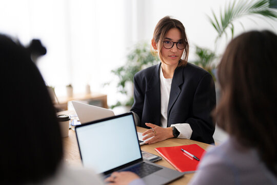 Young businesswoman in glasses engages in a focused discussion during a modern office meeting. Laptops and notes are on the table, indicating a productive work session.