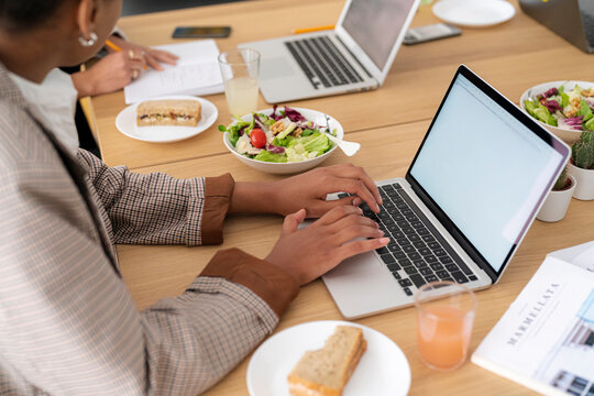 Two people work at a wooden table. One types on a laptop, another writes in a notebook. Healthy lunch items like salad and sandwiches are on the table, showing a productive session.