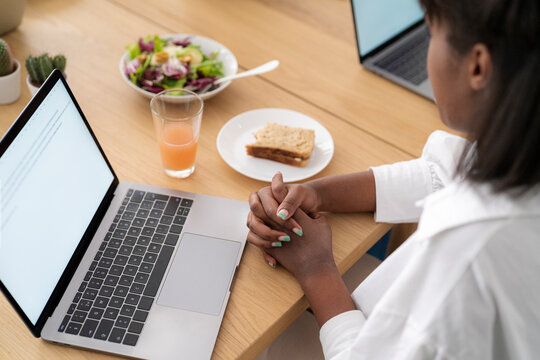 A woman sits at a wooden desk, focused on her laptop. A healthy lunch of salad, a sandwich, and juice is ready beside her. Her hands are clasped in thought.