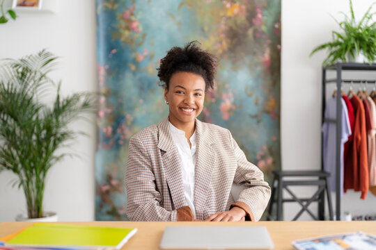 A smiling young Black woman sits at a desk in a bright office. She wears a plaid blazer, looking confidently at the camera. Her workspace includes a laptop and colorful folders.