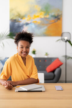A smiling young Black woman sits at a desk in a modern home office. She has a laptop, notebook, and phone on the wooden surface, ready for work or study.