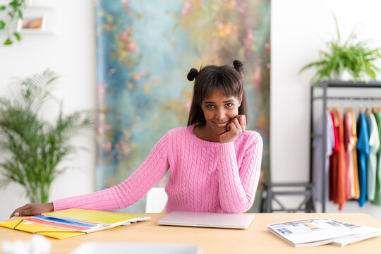 A smiling young woman with dark skin and hair in buns sits at a desk with a laptop and folders. She wears a pink sweater, looking at the camera. A colorful background is behind her.